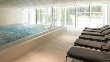 Ein Innenpool mit großen Glaswänden, gefliestem Boden und Liegestühlen.