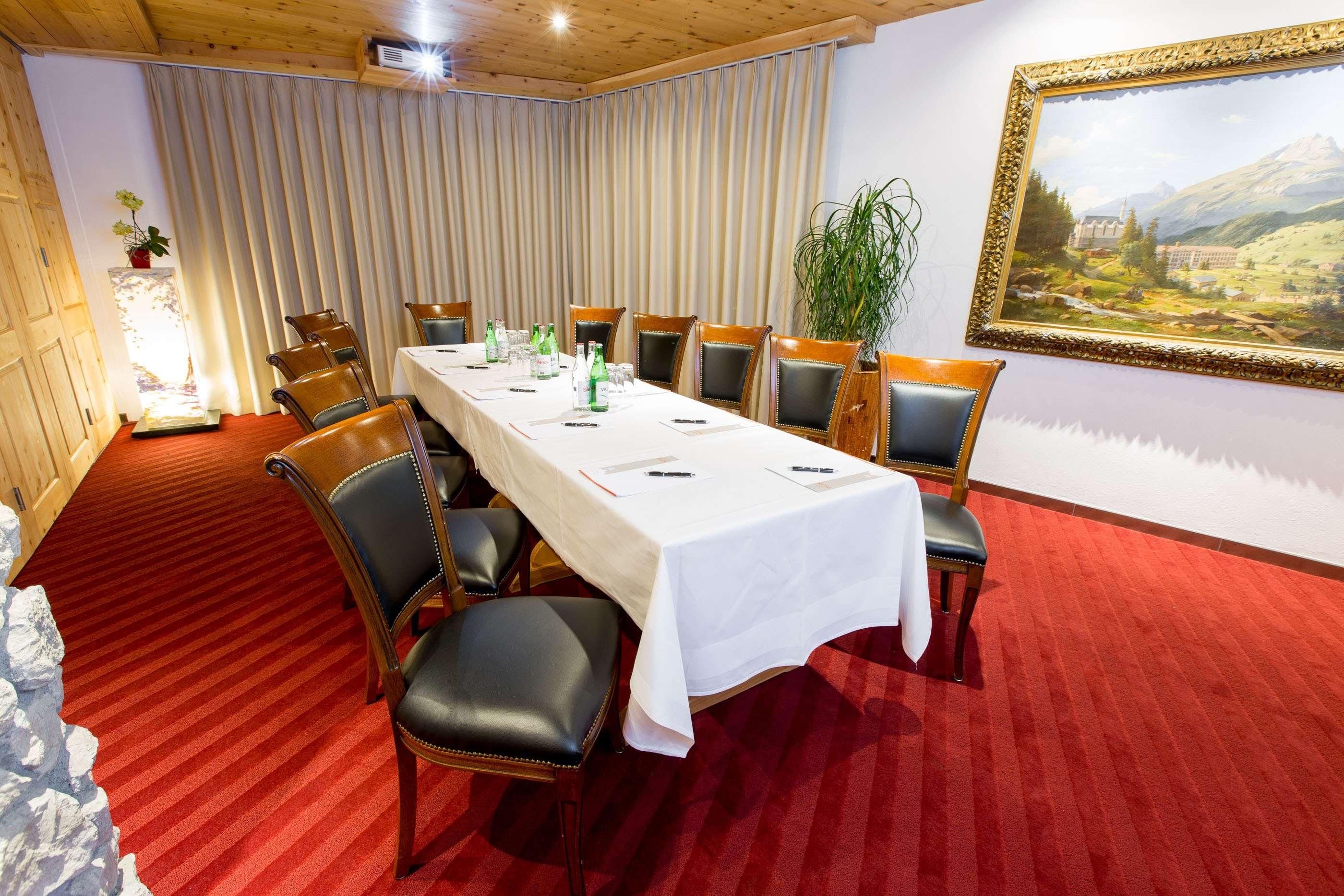 A conference room with a long table, chairs, a red carpet, a painting, and a plant.