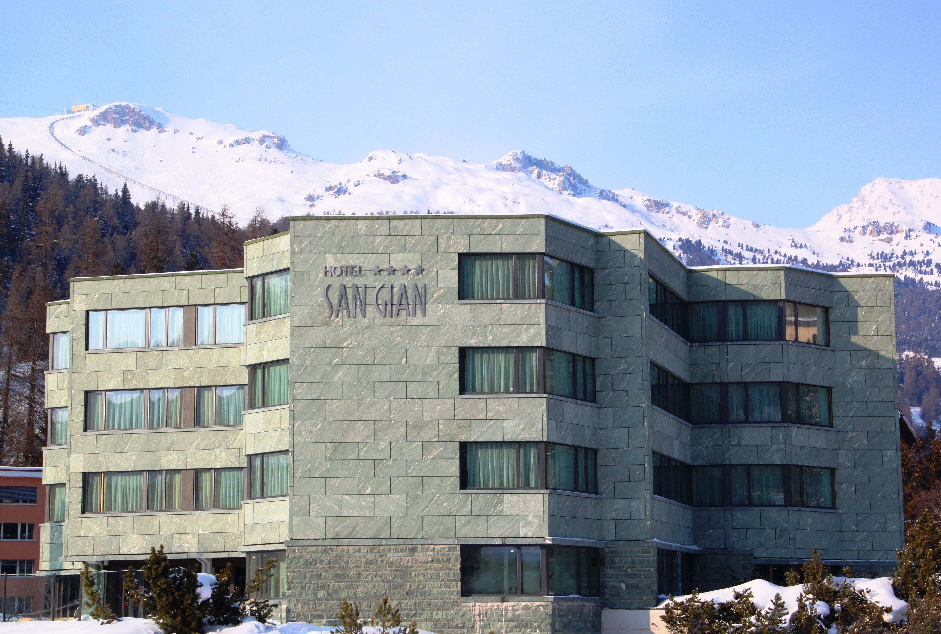 A modern hotel building with a stone facade, multiple windows, and a view of snow-capped mountains in the background.