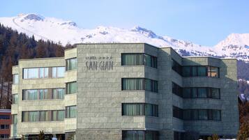 A modern hotel building with a stone facade, multiple windows, and a view of snow-capped mountains in the background.