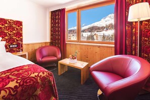 A hotel room with a bed, a red leather chair, a wooden table with a fruit arrangement, and a window with a mountain view.
