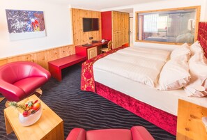 A hotel room with a bed, a desk, a red chair, a small table with fruit, and a mirror. - TOP Hotel San Gian St. Moritz (St. Moritz)