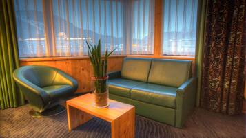 A room with a green sofa, a green chair, a wooden coffee table, and a view of a cityscape through large windows.