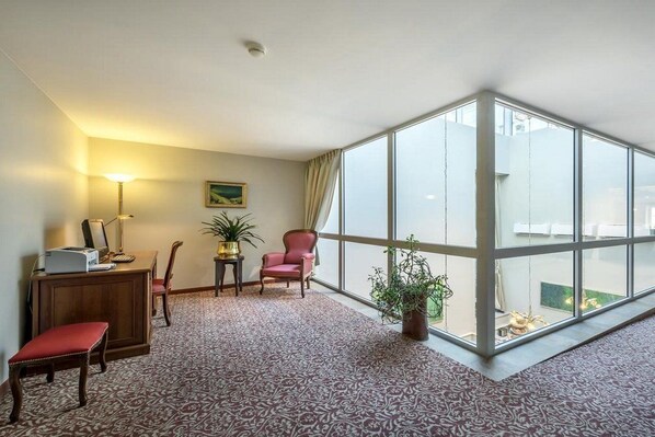 A hotel room with a large window, a desk with a lamp, a chair, a small table with a plant, and a patterned carpet. - Ratonda Centrum Hotel (Vilnius)
