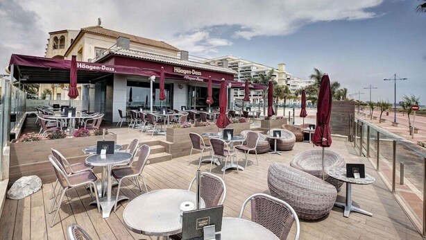 Ein Sitzbereich im Freien mit Tischen und Stühlen, eine überdachte Terrasse und ein Gebäude mit der Beschriftung "Häagen-Dazs".