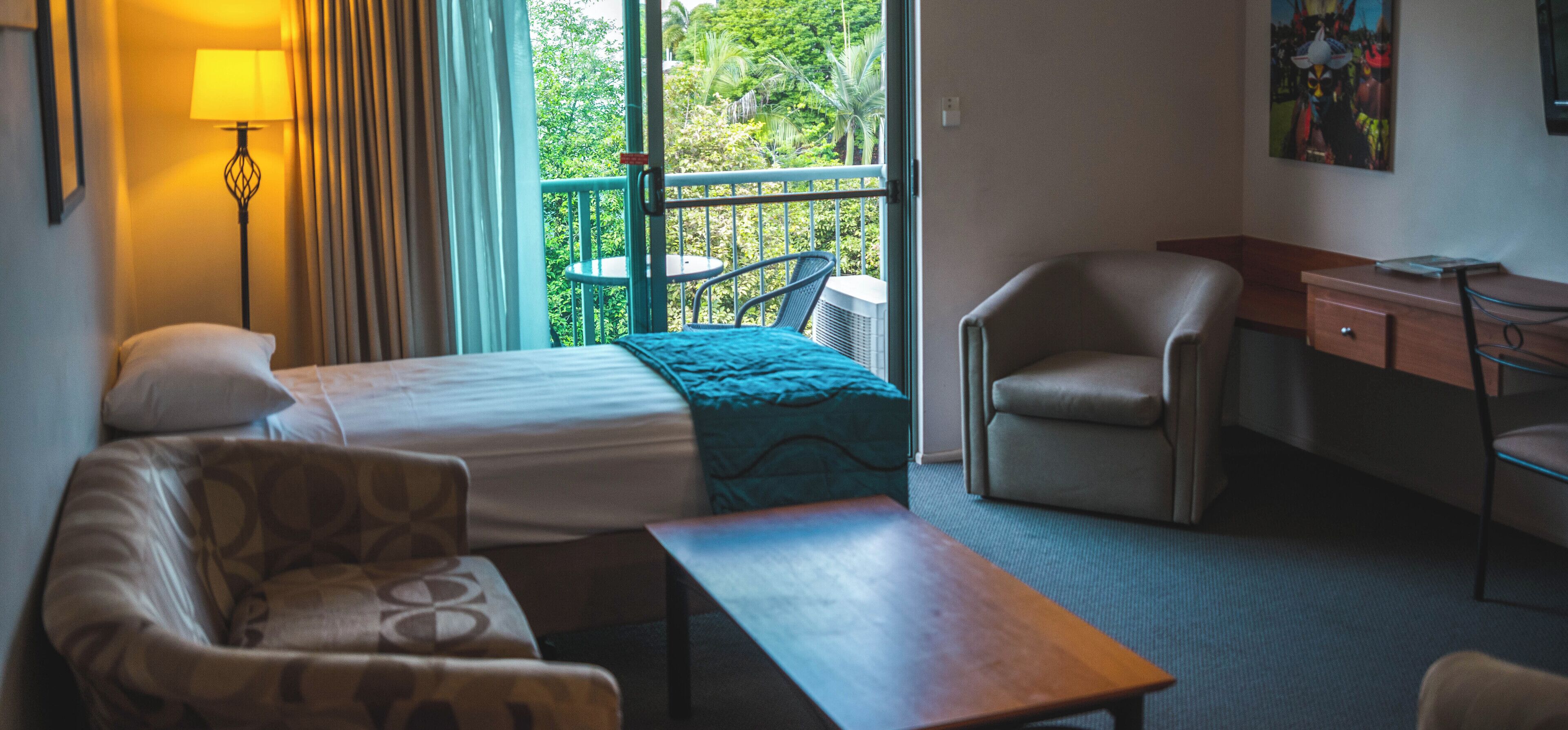 Habitación de hotel con cama, escritorio, silla, mesita pequeña y balcón con vista a zonas verdes.