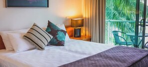 A hotel room with a bed, pillows, a nightstand with a lamp, and a view of greenery through a window. - Heritage Cairns (Cairns)