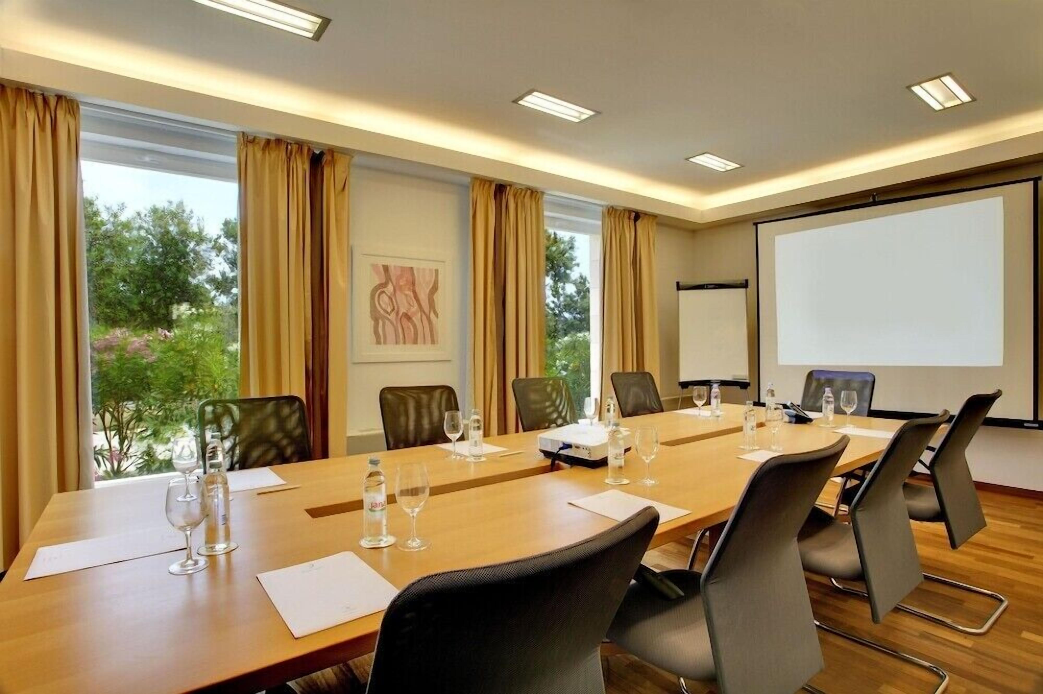 Un salón de conferencias con una larga mesa, sillas, una pantalla de proyector y vistas a zonas verdes.