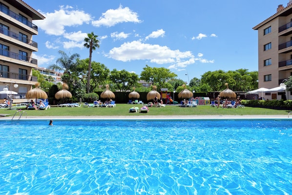Una piscina con sillones de descanso y sombrillas, rodeada de edificios y árboles.