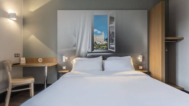 A hotel room with a bed, a desk, a chair, and a view of a building through a window. - B&B Hotel Orange (Orange)