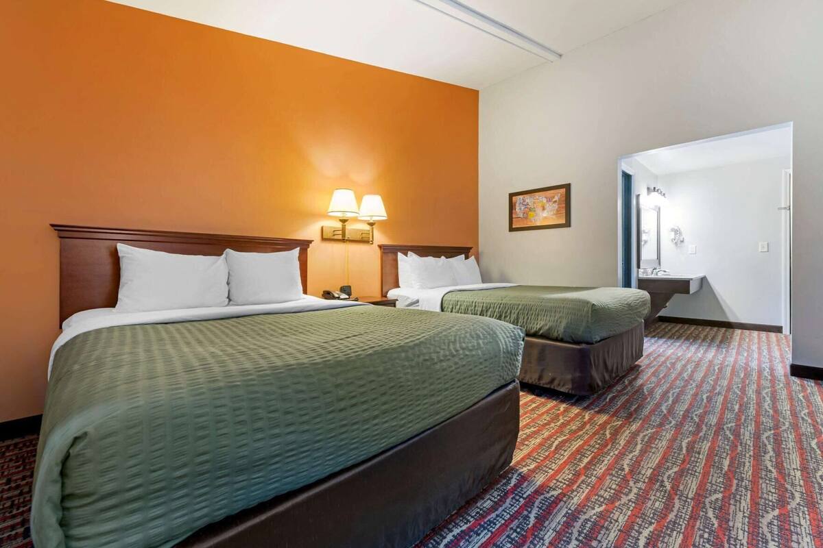 Una habitación de hotel con dos camas, una mesita de noche, una lámpara, un cuadro y un baño visible a través de una puerta abierta.