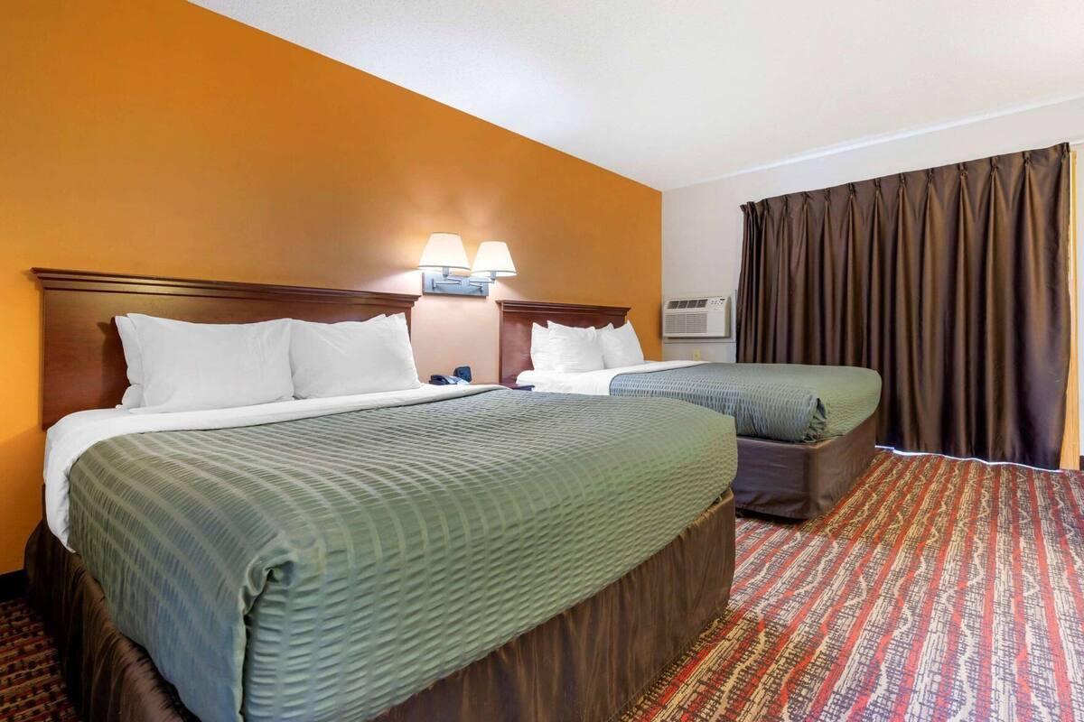 Habitación de hotel con dos camas, un ventanal con cortinas, aire acondicionado de pared y alfombra estampada.