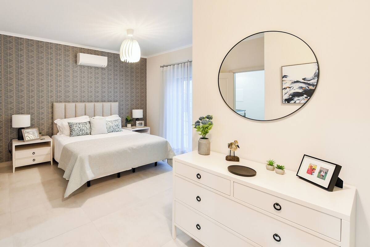A modern bedroom with a large bed, bedside tables, a dresser, a mirror, and a patterned wallpaper.