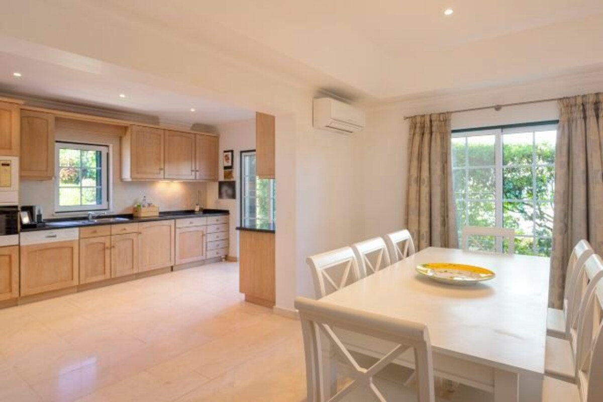 A modern kitchen with wooden cabinets, a dining table with chairs, and a large window with curtains.