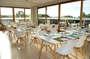 A spacious dining area with multiple tables and chairs, large windows offering a view of the outdoors.