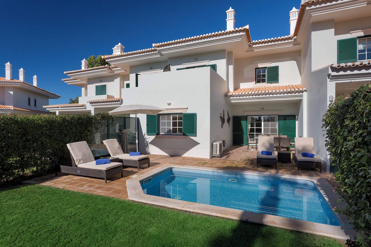 A poolside area with lounge chairs, a small pool, and a white building with green shutters.