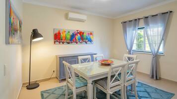 Uma área de refeições com mesa de vidro, cadeiras brancas, pintura de um papagaio colorido, um abajur e uma janela com cortinas.