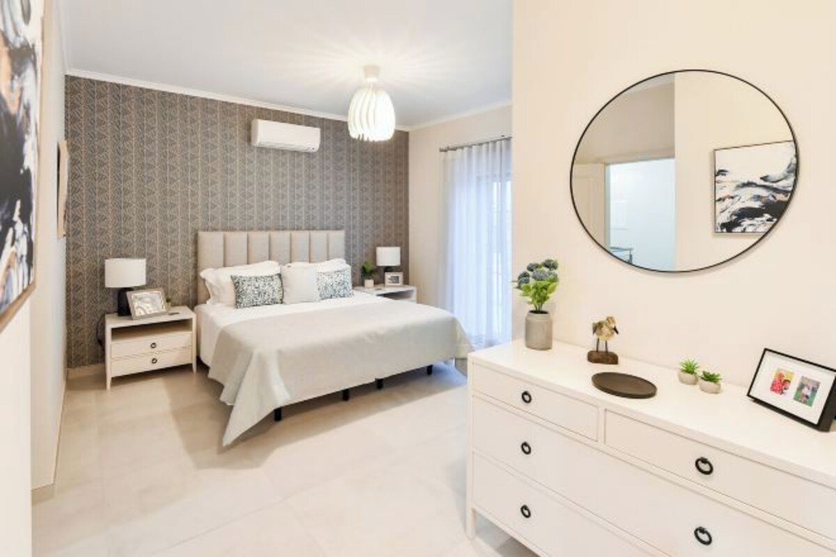 A modern bedroom with a large bed, bedside tables, a round mirror, and a dresser.