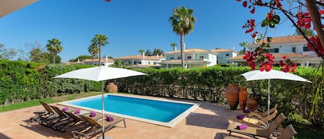 A swimming pool with lounge chairs and umbrellas, surrounded by greenery and a view of a large house.