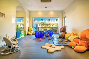 Zona de juegos infantiles con asientos cómodos, juguetes y vistas a un edificio a través de un ventanal.