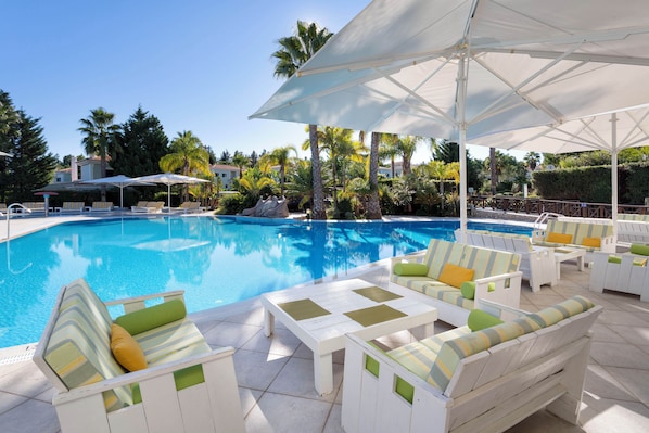 A pool area with lounge chairs and umbrellas, surrounded by palm trees and a clear blue sky. - Martinhal Quinta Do Lago Family Resort (Loulé)