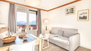 A cozy living room with a sofa, a dining table with chairs, a small side table, and a view of mountains through a large window. - Residence Les Bergers (Huez)