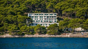 Un hotel de diverses plantes amb terrassa, envoltat de vegetació arbustiva i arbres frondosos, amb vista a una massa d'aigua.