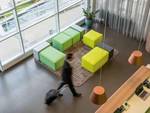 Una hall moderna con ampie vetrate, un'area salotto con cuscini verdi e gialli, una pianta in vaso e una reception con un laptop.