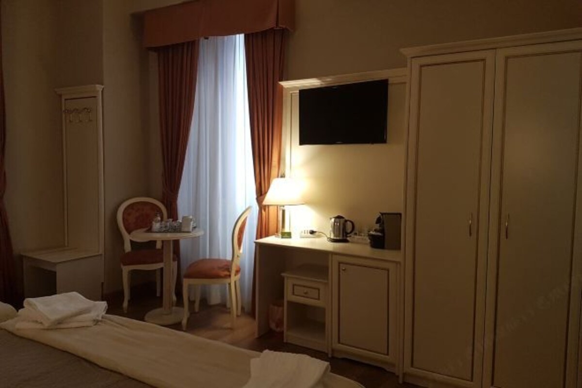 A hotel room with a bed, a desk with a lamp, a chair, a wardrobe, and a television on the wall.