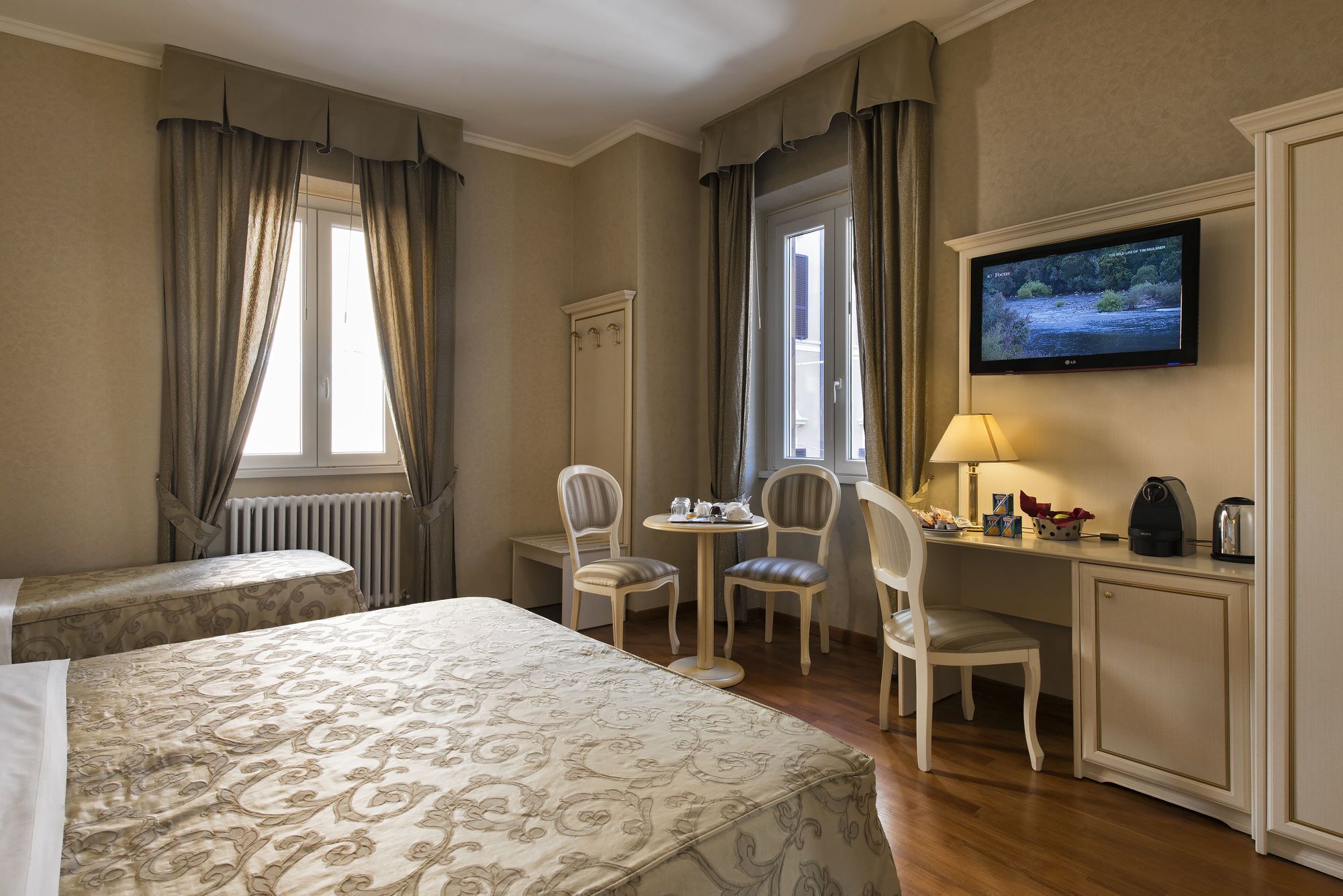 A hotel room with a bed, two chairs, a small table, a desk, a television, and a radiator.