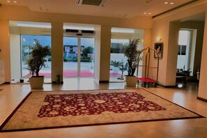 Een hotel lobby met een groot rood tapijt, potplanten en uitzicht op de straat.