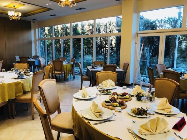 A dining area with round tables, chairs, and a view of trees through large windows. - Grand White City Hotel (Berat)