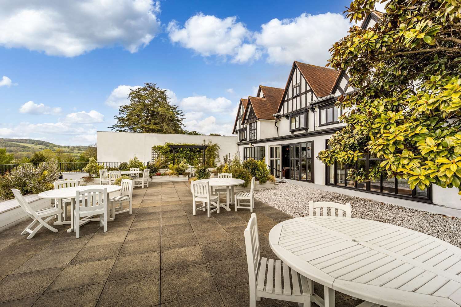 Un patio con mesas y sillas blancas, un jardín con diversas plantas y un edificio de estilo arquitectónico tradicional.