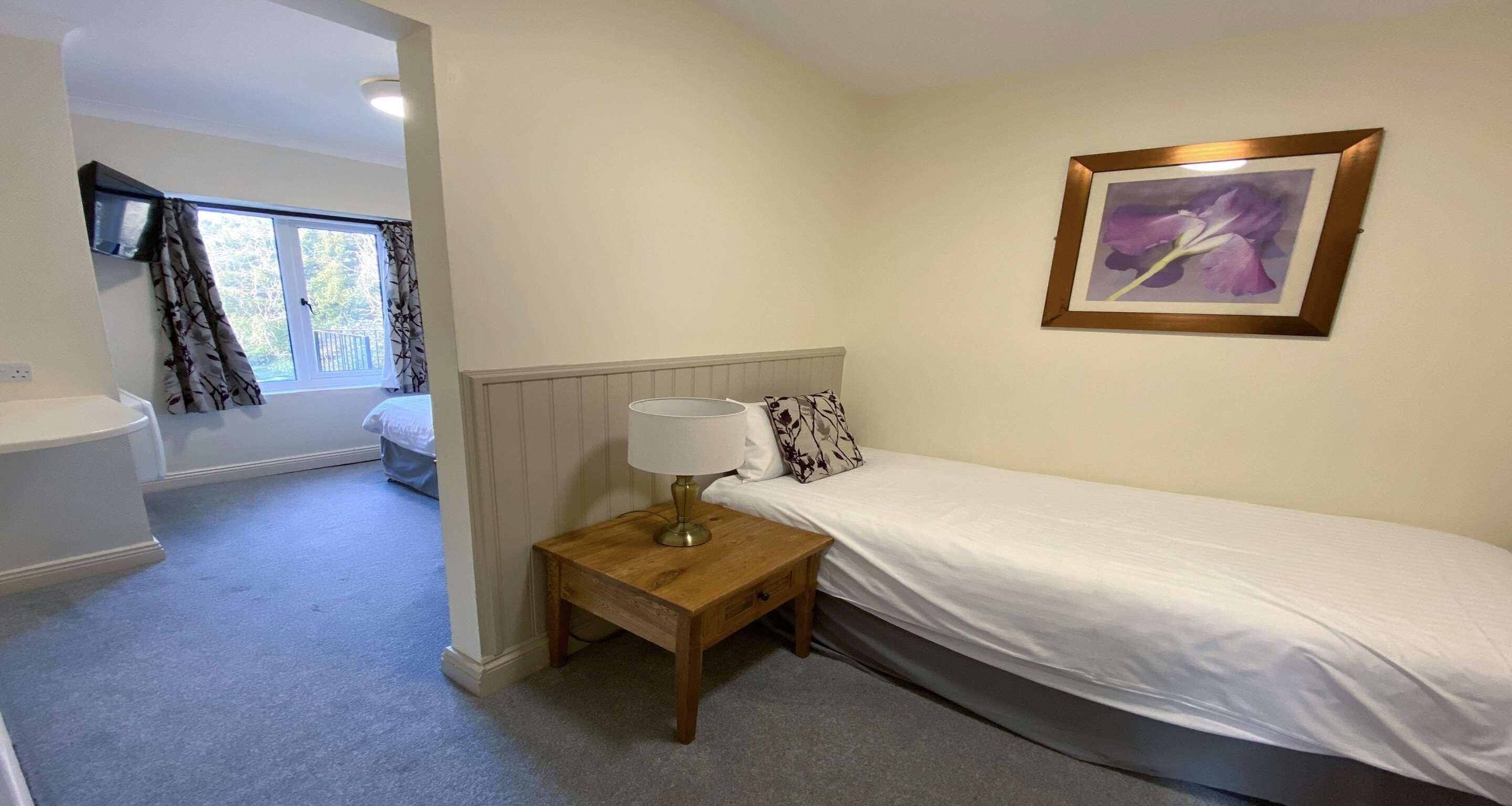 Une chambre avec un lit simple, une table de chevet avec une lampe, un tableau encadré au mur et une fenêtre avec des rideaux.