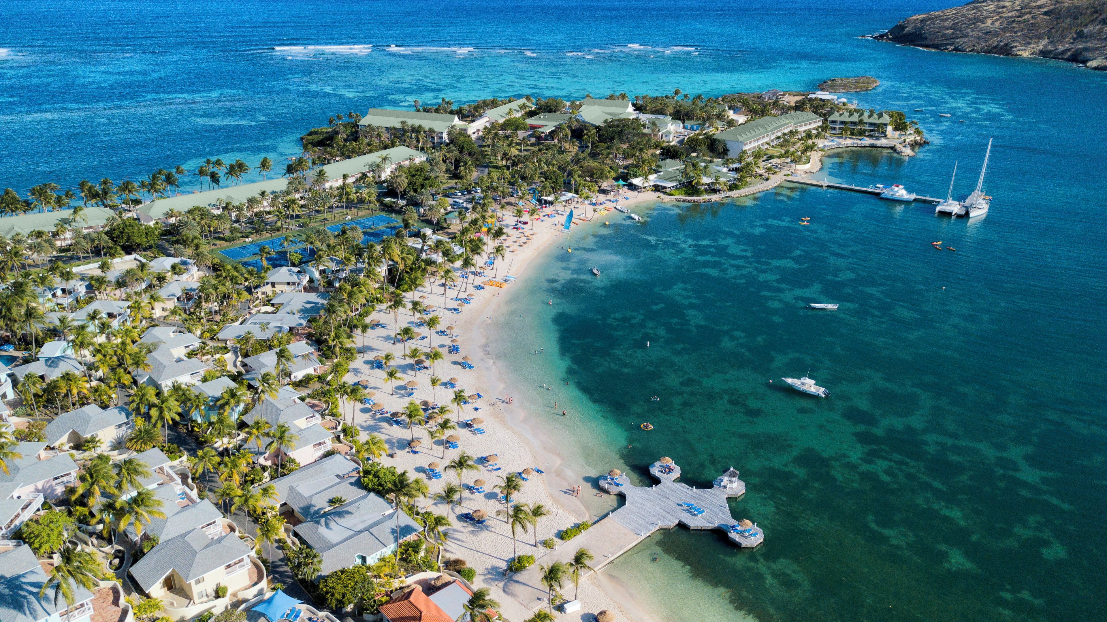 An aerial view of a coastal resort with multiple buildings, a beach, and clear blue waters.
