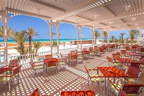Un espace extérieur abrité, doté de tables et de chaises, offrant une vue sur une plage et l’océan.