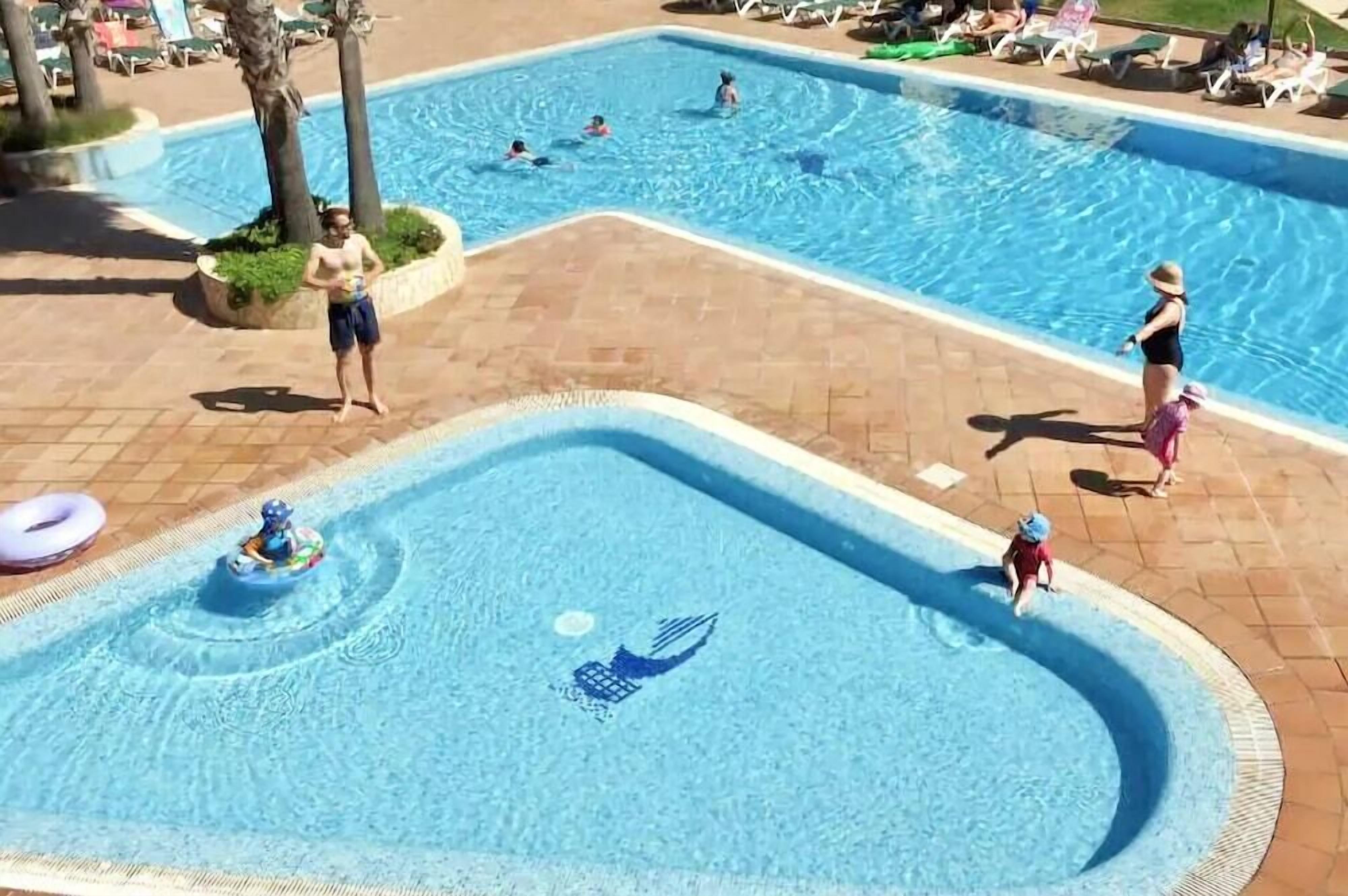 Zona de piscina con varias piscinas, sillas de descanso y una pequeña área de juegos acuáticos para niños.