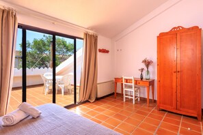 A room with a bed, a desk, a chair, a wardrobe, and a sliding glass door leading to an outdoor area with a table and chairs.