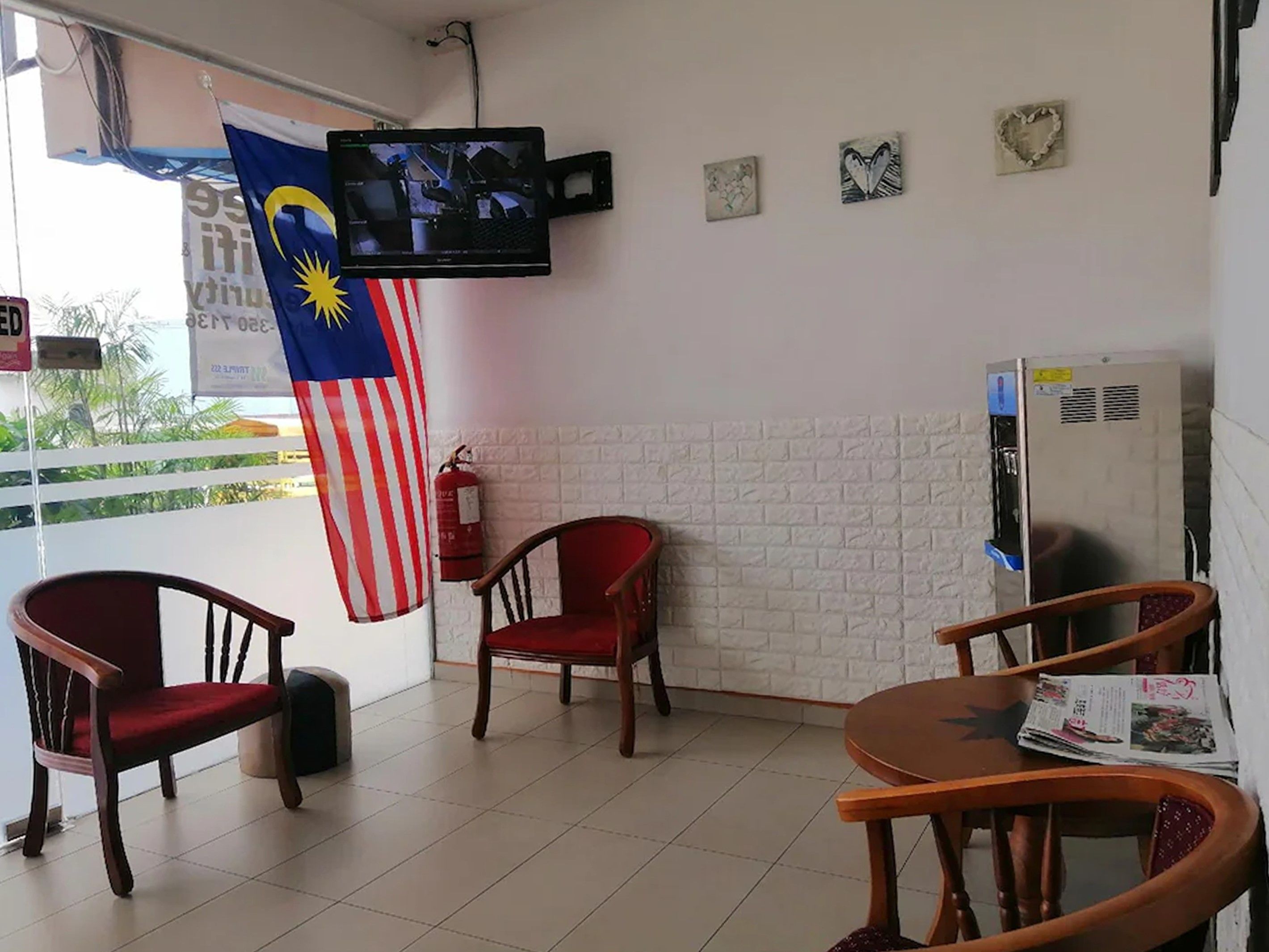 A room with a television mounted on the wall, a fire extinguisher, and a flag. There are wooden chairs and a table with newspapers.