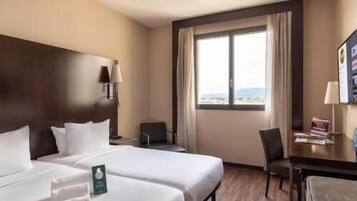 A hotel room with two beds, a desk, a chair, a TV, and a window with a view.
