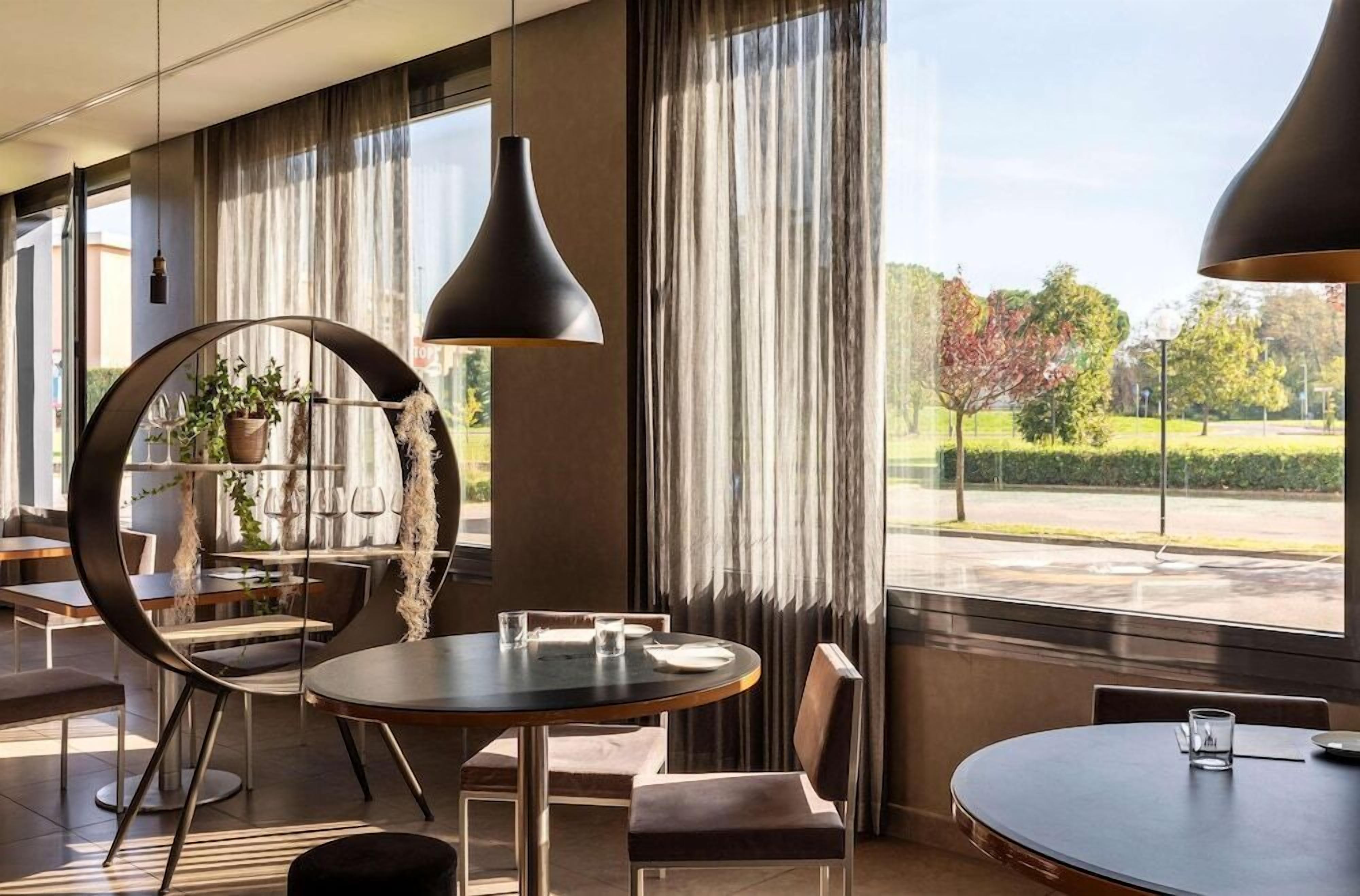 Un comedor moderno con mesas y sillas redondas, ventanas amplias con cortinas y lámparas colgantes.