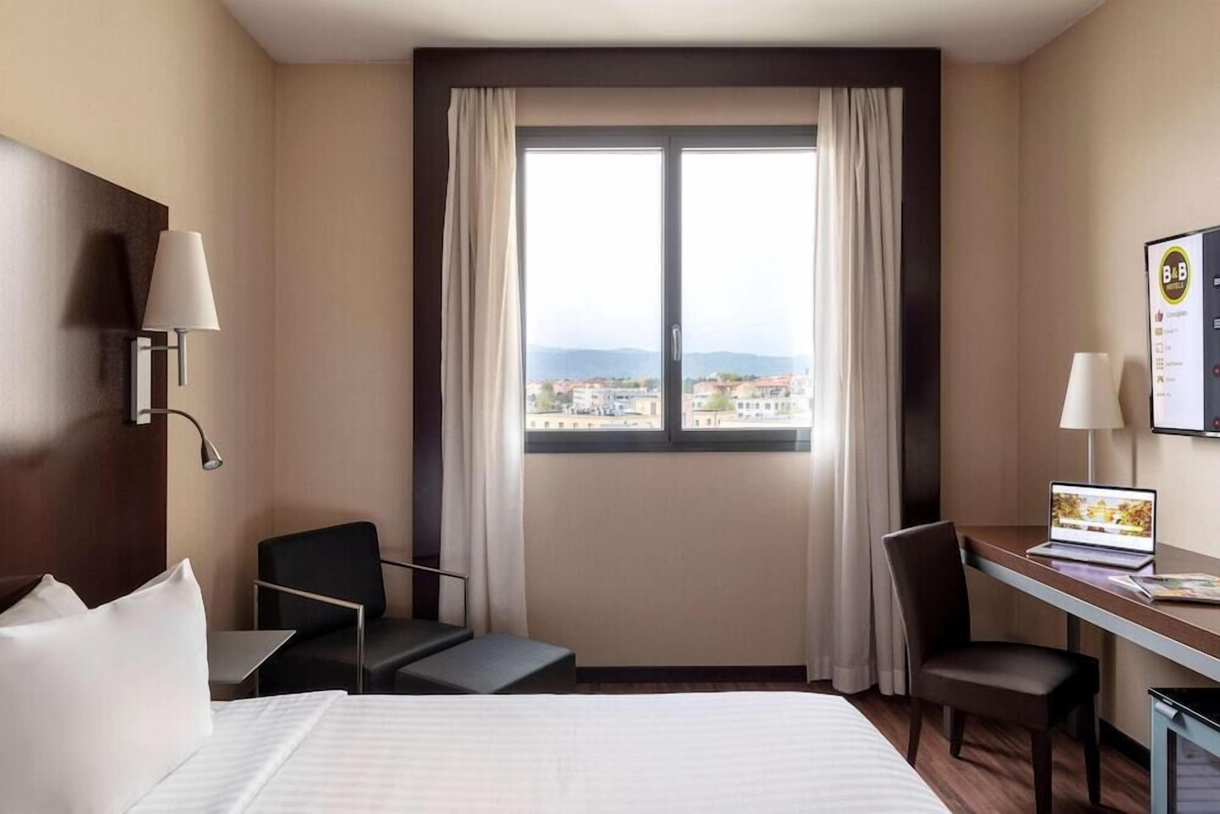 A hotel room with a bed, two chairs, a desk, a laptop, and a window with curtains.