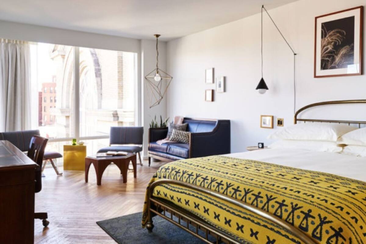 Ett hotellrum med en säng, ett skrivbord, en stol, en soffa och ett fönster med utsikt över byggnader.