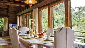 A dining area with a wooden table set for breakfast, surrounded by chairs, large windows, and a view of the forest. - Paraíso na Montanha House Boutique (Campos do Jordão)