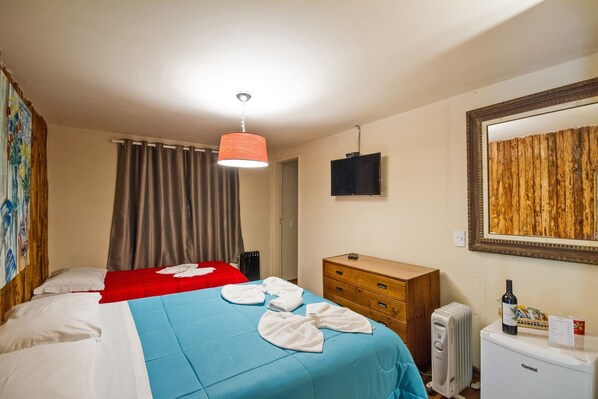 Habitación de hotel con dos camas, una colcha roja y otra azul, un aparador de madera, un radiador, un televisor y un espejo en la pared.