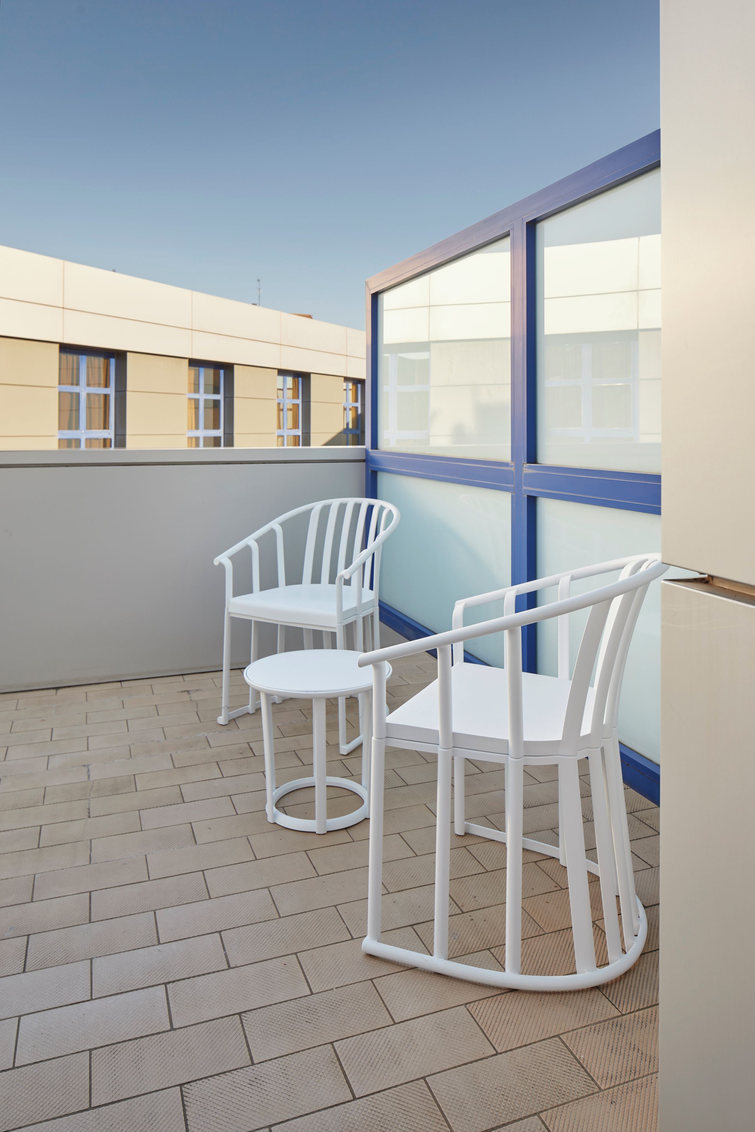 A balcony with two white chairs and a table, a large window, and a tiled floor.