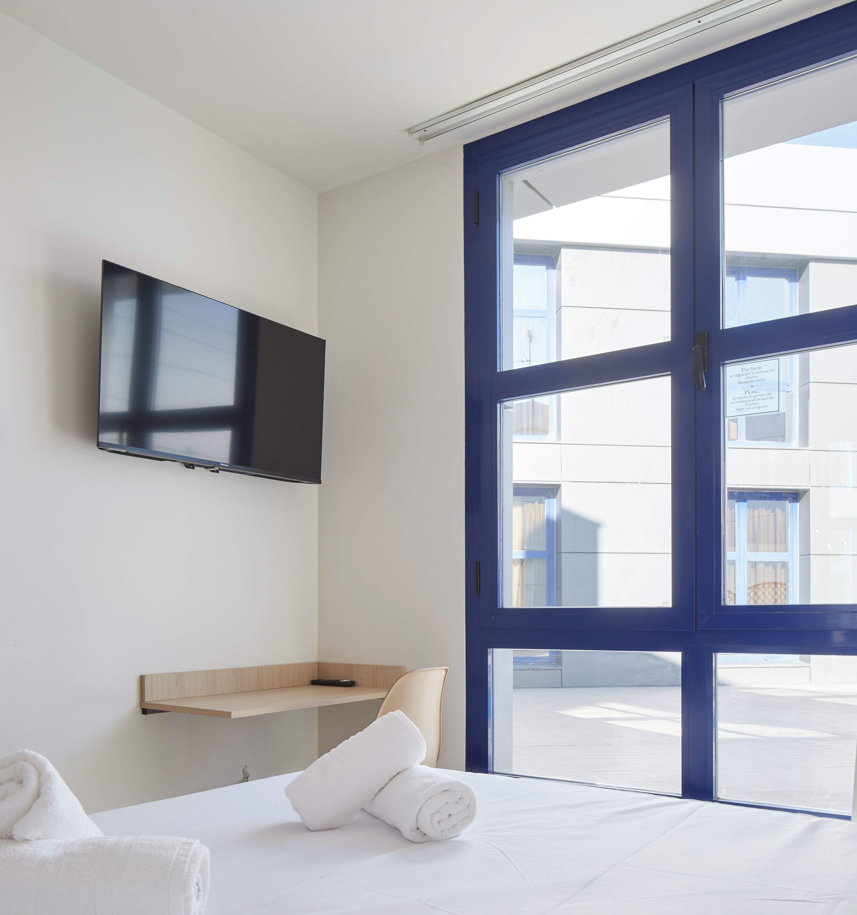 A hotel room with a bed, a TV mounted on the wall, a window with a view of buildings, and a wooden shelf.