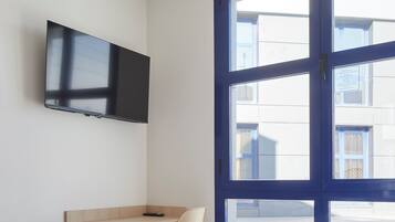A hotel room with a bed, a TV mounted on the wall, a window with a view of buildings, and a wooden shelf.