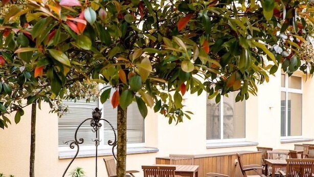 Uma área de estar ao ar livre com mesas e cadeiras de madeira, rodeada por vegetação e um suporte de metal decorativo.