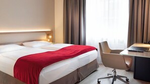 Quarto de hotel com cama, escrivaninha, cadeira e janela com cortinas.
